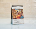 A spiral-bound New Testament desk calendar standing upright on a light marble surface against a beige tiled wall. The front cover shows a classical painting with figures in warm tones and text reading 'NEW TESTAMENT' and 'Come Follow Me 2027'.
