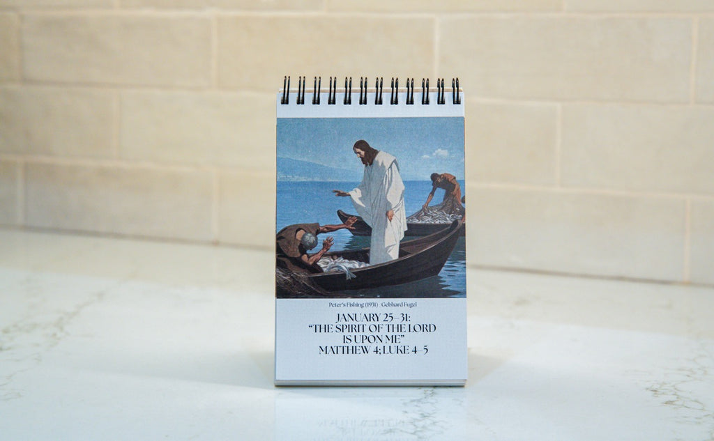 A spiral-bound standing calendar or notebook with a printed illustration of Jesus in a boat and a Bible verse at the bottom, photographed upright on a light marble-like surface against a beige tiled wall.