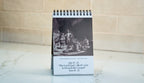 A spiral-bound desk calendar or print standing upright on a light countertop, with a black-and-white historical scene on the upper portion and printed text on the lower white panel. The background is a softly blurred beige tiled wall.