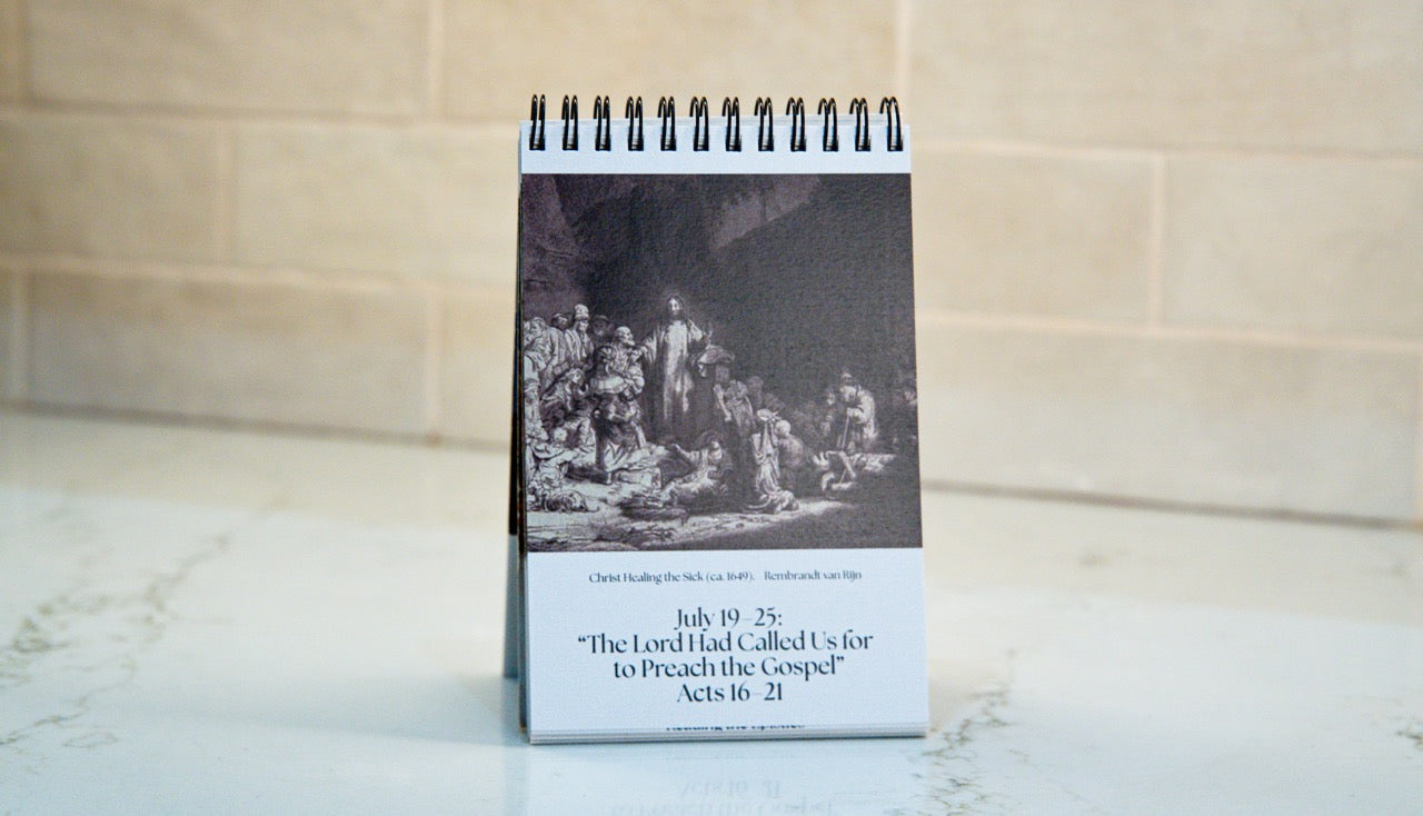 A spiral-bound desk calendar or print standing upright on a light countertop, with a black-and-white historical scene on the upper portion and printed text on the lower white panel. The background is a softly blurred beige tiled wall.