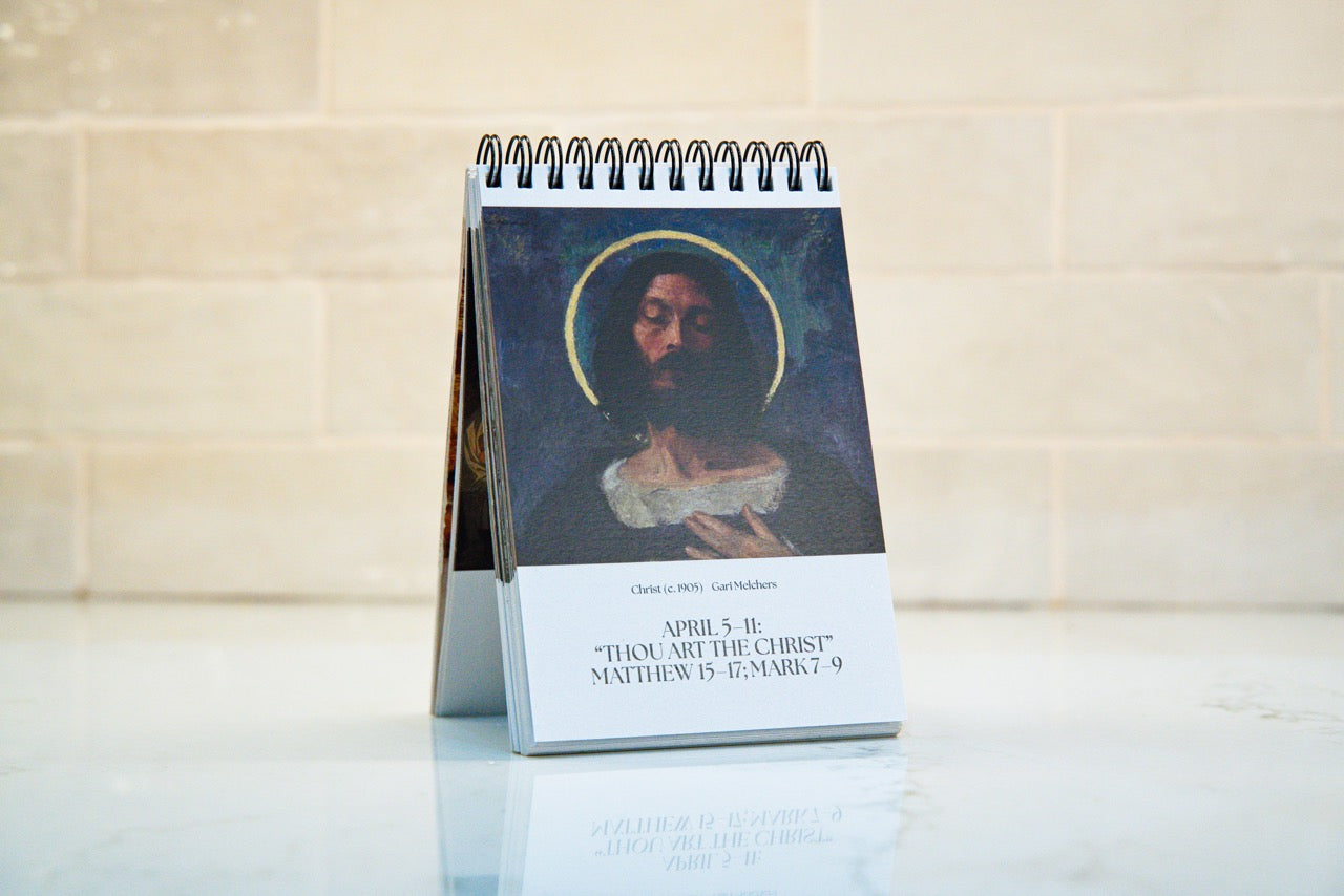 A spiral-bound standing Bible calendar featuring a religious illustration of Jesus with a halo and the text 'THOU ART THE CHRIST' and 'APRIL 5th, 2026 MATTHEW 5:3-9', displayed on a reflective white surface against a beige stone wall.