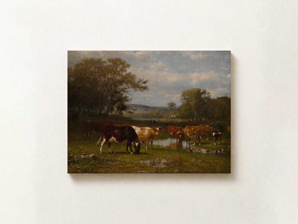 A rectangular canvas print of a classic landscape painting showing cows grazing and drinking from a pond in a grassy field under a cloudy sky, mounted on a light beige wall.