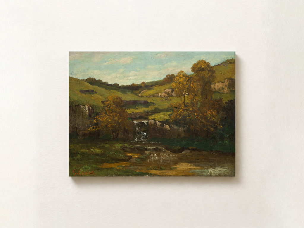 A landscape oil painting on canvas, depicting rolling green hills, trees with autumnal yellow and orange foliage, a small waterfall, and a river under a pale blue sky, shown hanging on a textured off-white wall.