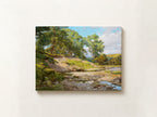 A horizontal landscape painting of a rocky stream with green trees and grassy banks under a blue sky, displayed on a light beige wall.