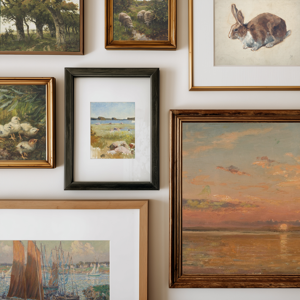 A gallery wall display of multiple framed art prints and paintings in various frames, including landscapes, a rabbit sketch, ducks, and a sunset seascape, arranged on a white wall.