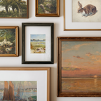 A gallery wall display of multiple framed art prints and paintings in various frames, including landscapes, a rabbit sketch, ducks, and a sunset seascape, arranged on a white wall.