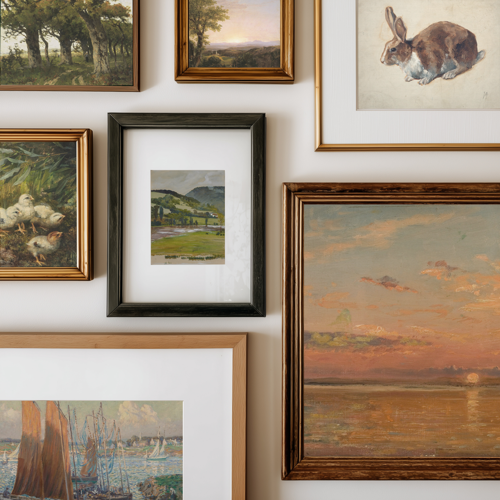 A gallery wall display of multiple framed paintings and art prints in different styles and colors, including landscapes, a rabbit sketch, ducks, and a sunset scene.