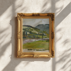 A framed landscape painting of rolling green hills and a river, set in an ornate gold frame and mounted on a textured white wall with soft shadow patterns.