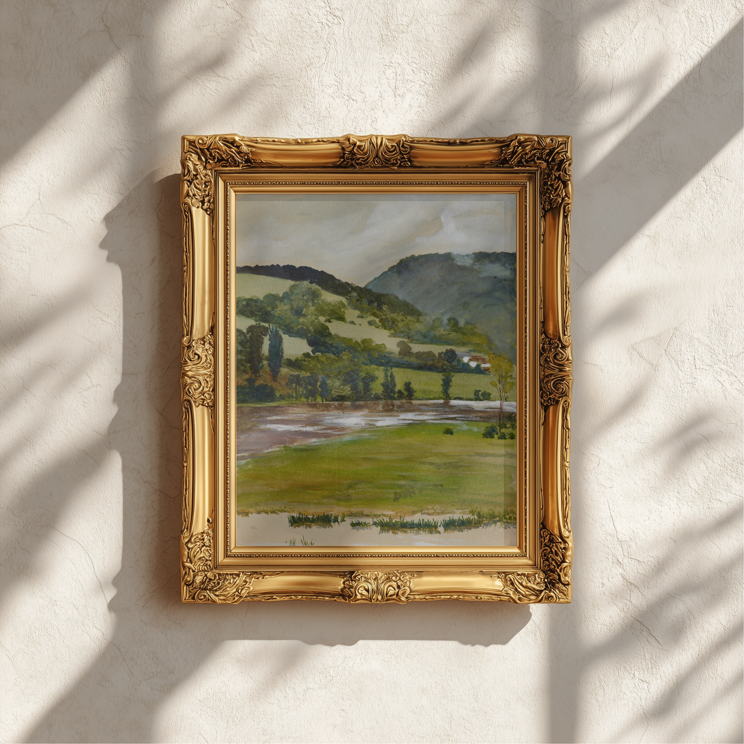 A framed landscape painting of rolling green hills and a river, set in an ornate gold frame and mounted on a textured white wall with soft shadow patterns.