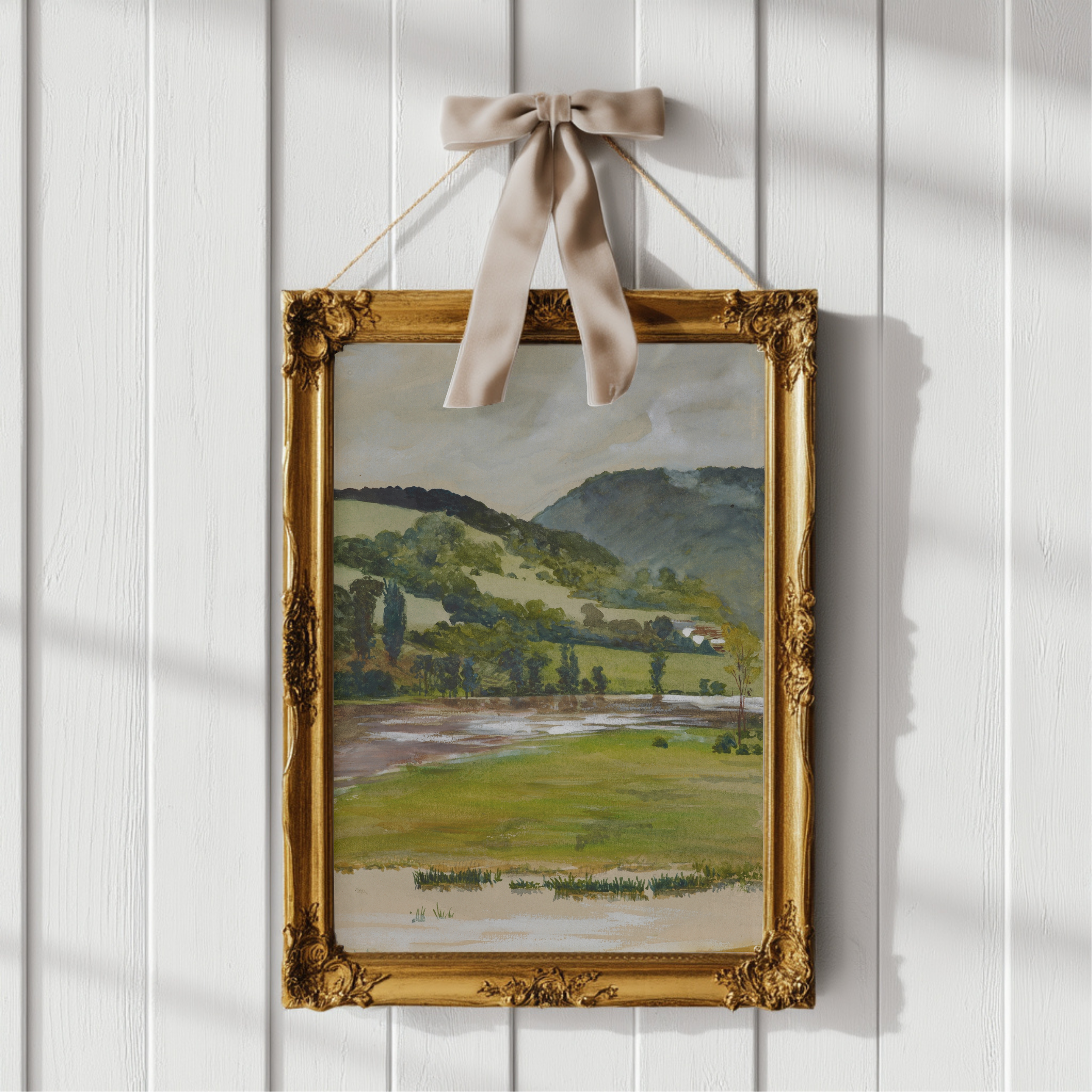 A framed landscape painting hanging on a white paneled wall. The artwork depicts green fields, trees, hills, and a pale sky, enclosed in an ornate gold frame with a beige ribbon bow at the top.
