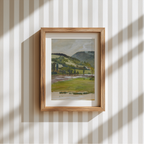 A framed landscape art print featuring green fields, trees, and distant hills, displayed on a cream and white vertically striped wallpaper background with soft window shadow patterns.