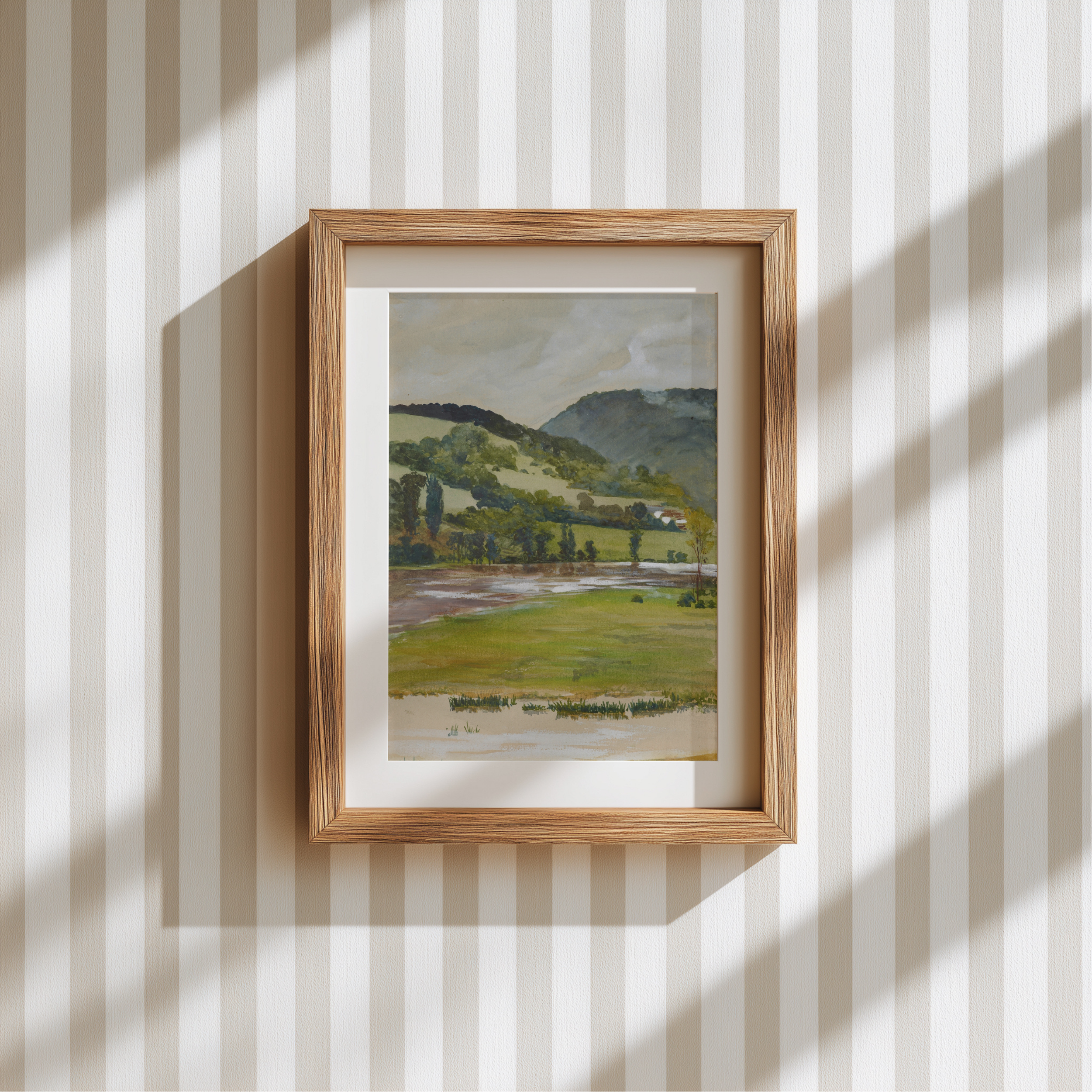 A framed landscape art print featuring green fields, trees, and distant hills, displayed on a cream and white vertically striped wallpaper background with soft window shadow patterns.