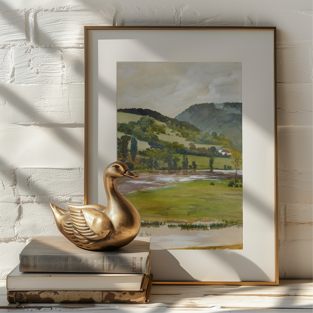 A framed landscape art print leaning against a white painted brick wall, styled with a gold-toned duck sculpture and stacked books in the foreground.