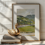 A framed landscape art print leaning against a white painted brick wall, styled with a gold-toned duck sculpture and stacked books in the foreground.