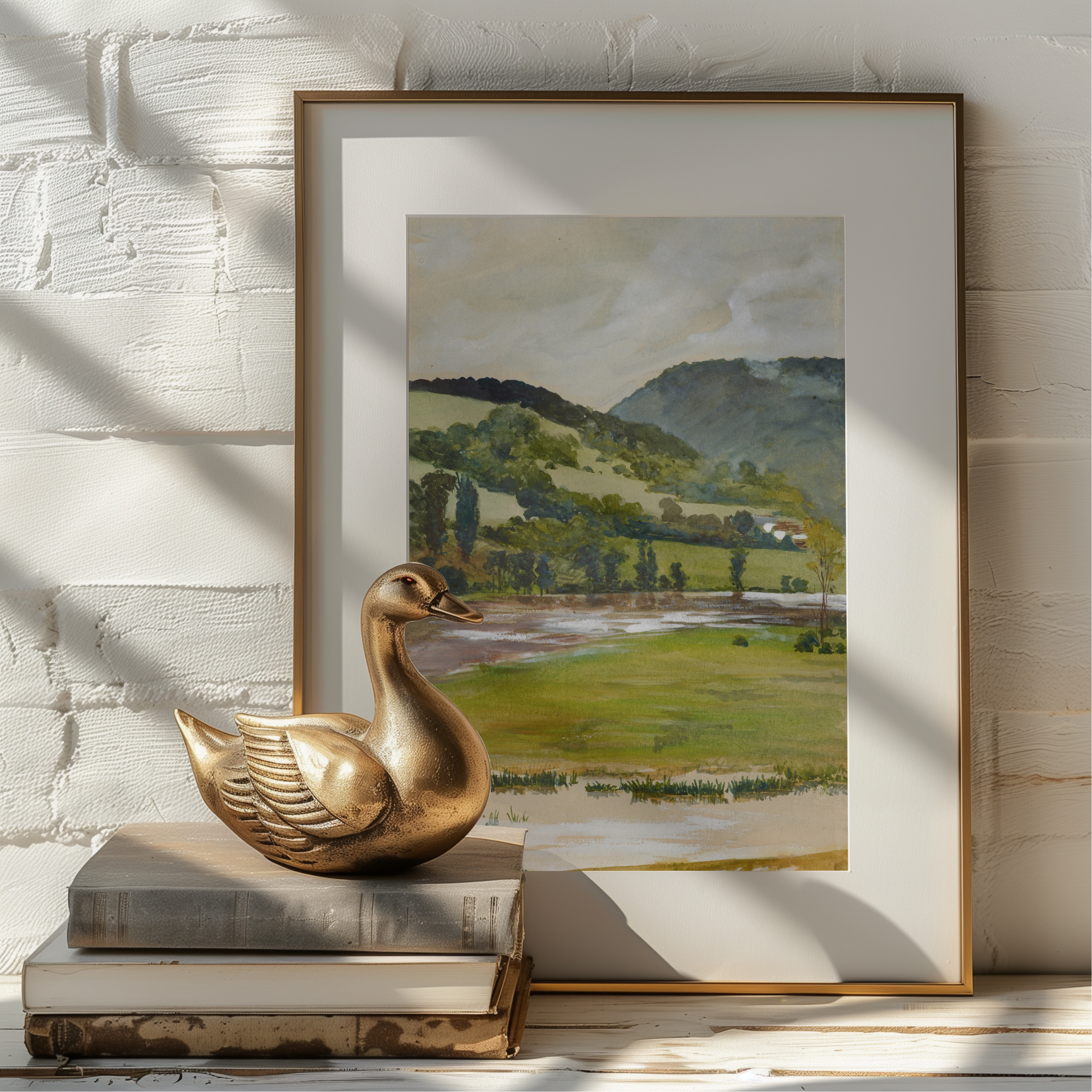 A framed landscape art print leaning against a white painted brick wall, styled with a gold-toned duck sculpture and stacked books in the foreground.