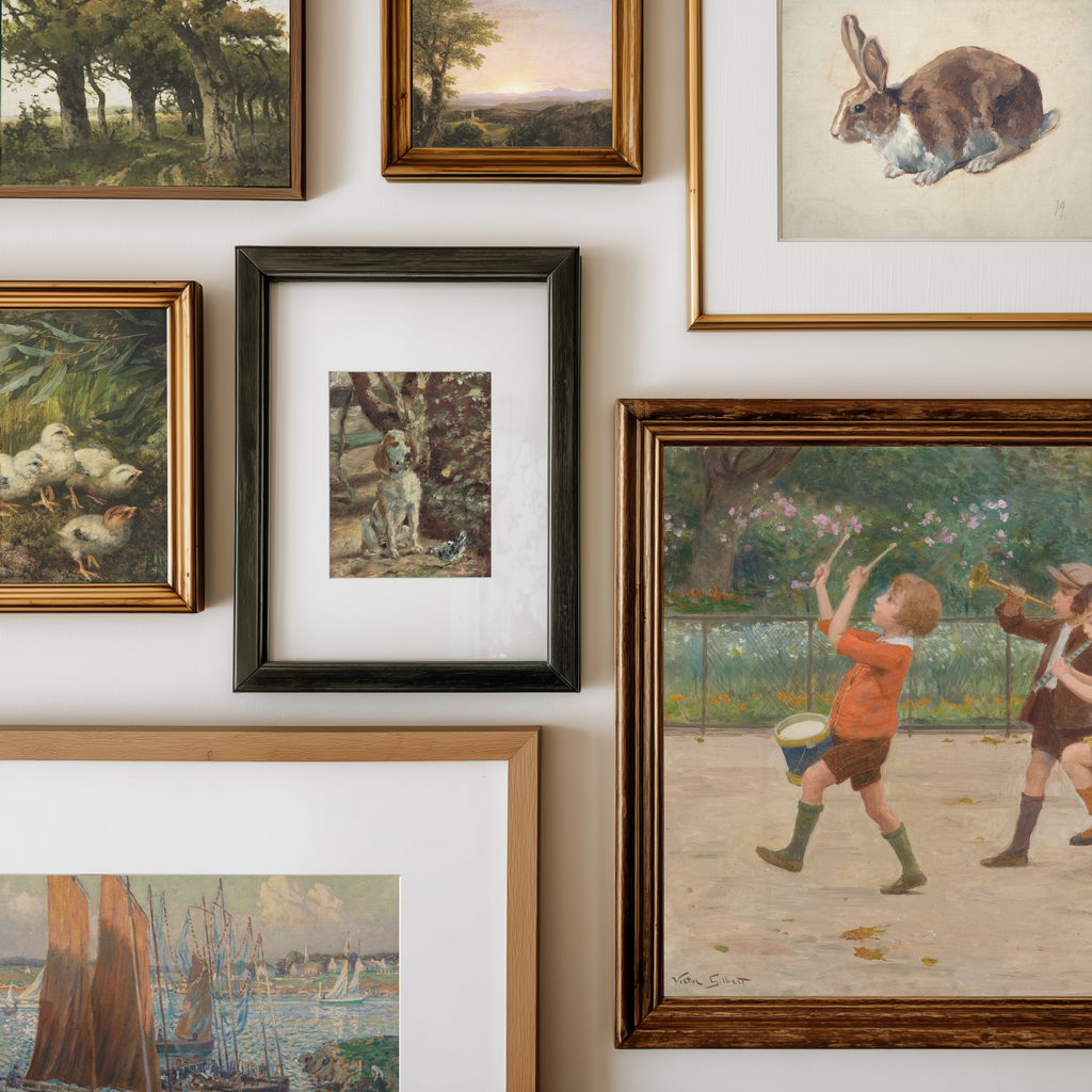 A gallery wall display of multiple framed art prints, including landscapes, a rabbit portrait, ducks, a dog, children playing, and a harbor scene, arranged on a white wall.