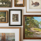 A gallery wall display of multiple framed art prints and paintings, including landscapes, a rabbit portrait, ducks, a dog, and a rabbit in a black frame, arranged on a white wall.
