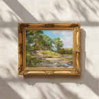 An ornate gold picture frame holding a landscape painting with green trees, a river, and blue sky, mounted on a textured white wall with dappled sunlight and shadows.