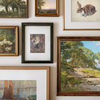 A gallery wall display of multiple framed art prints and paintings, including landscapes, a rabbit illustration, ducks, and a central dark-framed artwork, arranged on a white wall.