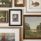 A gallery wall display of multiple framed art prints and paintings, including landscapes, a rabbit study, ducks, and a central dark-framed artwork, arranged on a white wall.
