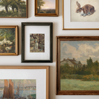 A gallery wall display of multiple framed art prints and paintings, including landscapes, a rabbit portrait, ducks, a dog, and a house scene, arranged on a white wall.