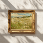 An ornate gold picture frame holding a landscape painting with green fields and a blue sea, mounted on a textured white wall with soft leaf shadows.