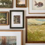 A gallery wall display of multiple framed art prints and paintings, including landscapes, a rabbit portrait, ducks, a dog, and a field scene, arranged on a white wall.