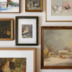 A gallery wall display of multiple framed paintings in different styles and colors, including winter landscapes, portraits, and religious scenes, arranged on a white wall.