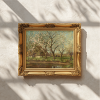 An ornate gold picture frame containing a landscape painting of blooming trees, mounted on a textured white wall with soft window shadow patterns.