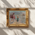 An ornate gold-framed painting depicting children playing on a beach, hung on a textured off-white wall with soft leaf shadows cast across it.