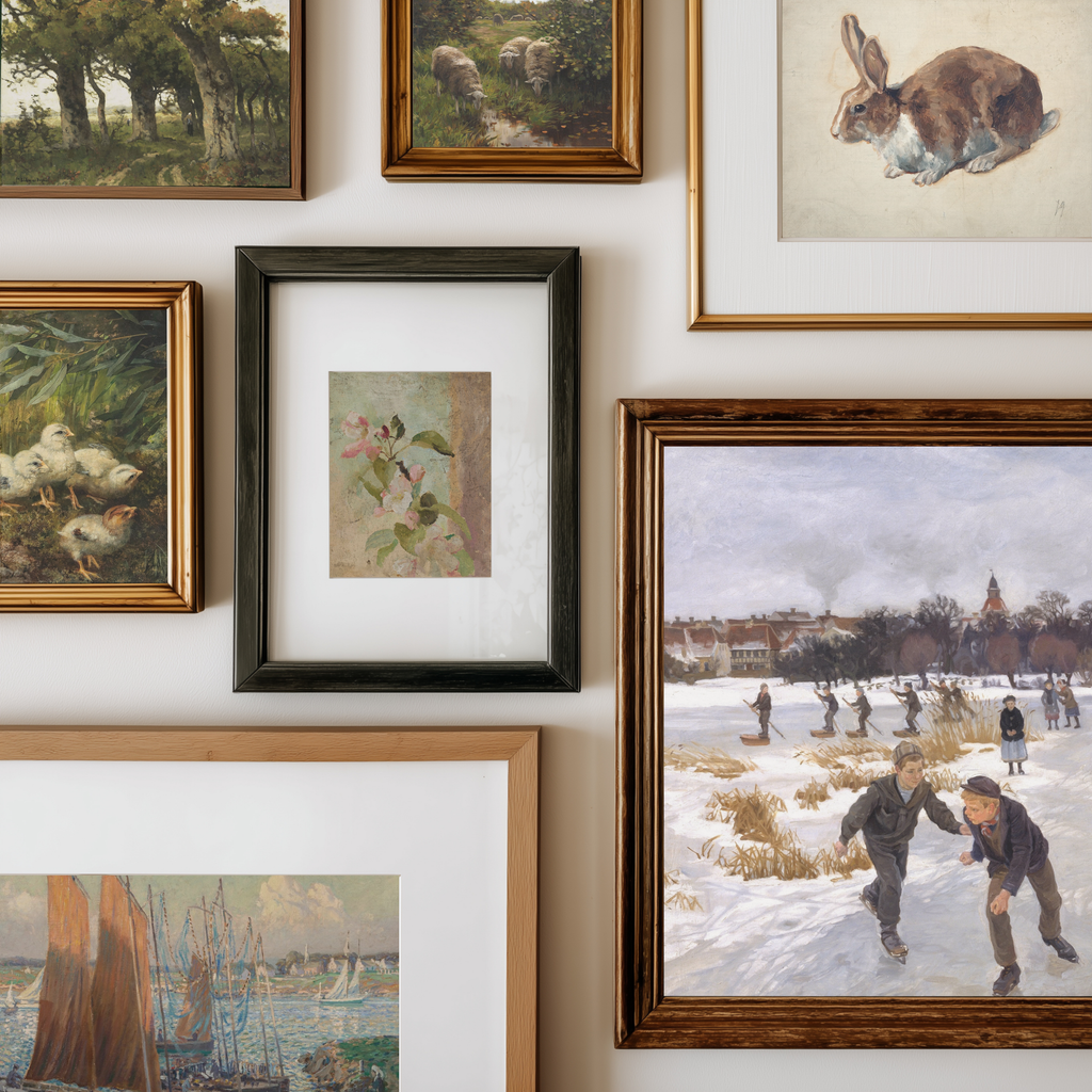A gallery wall display of multiple framed art prints and paintings in various frames, including landscapes, a rabbit illustration, ducks, a small floral painting, and a winter scene with people skating in the snow.