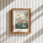 A framed art print depicting a seaside picnic scene with figures, a dog, and a harbor view, mounted on a cream and white vertically striped wallpaper background with soft window shadows.