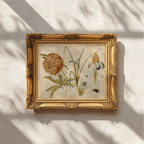 An ornate gold-framed botanical art print featuring a detailed illustration of a large orange-brown flower with green leaves, displayed on a textured off-white wall with soft window shadow patterns.