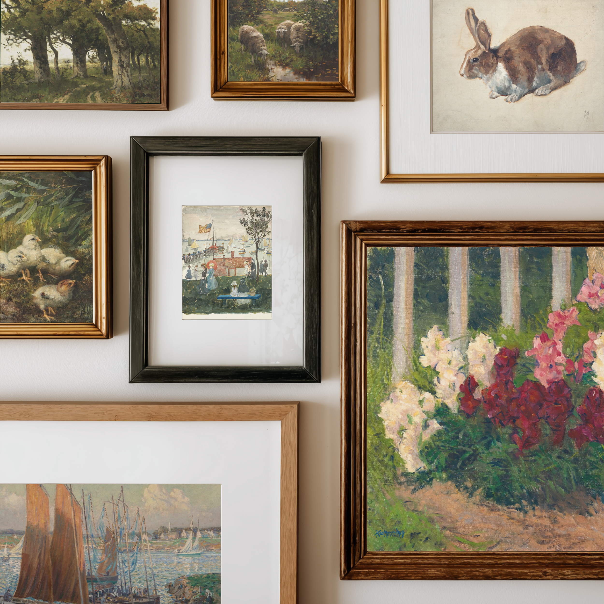 A gallery wall display of multiple framed art prints and paintings, including landscapes, a rabbit illustration, ducks, a small village scene, and a floral garden painting, arranged on a light wall.
