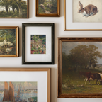 A gallery wall display of multiple framed art prints and paintings, including landscapes, animals, and a small floral scene, arranged on a white wall.
