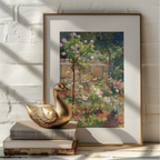 A framed art print depicting a lush, colorful garden with pink and white flowers under green foliage, styled on a white brick wall next to a gold-framed print, a gold swan figurine, and stacked books.