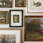 A gallery wall display of multiple framed art prints and paintings, including landscapes, animals, and a small floral scene, arranged on a white wall.