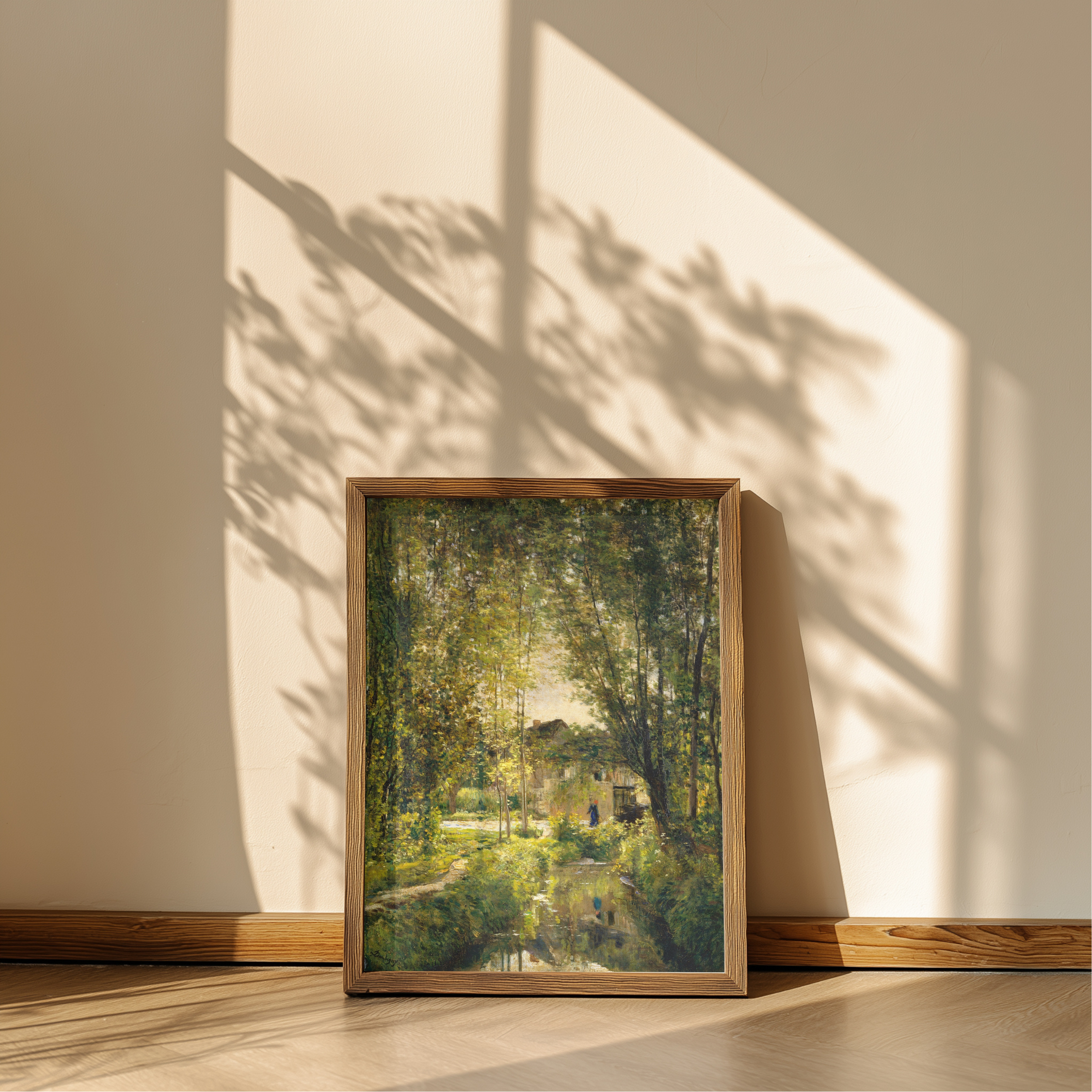 A framed landscape art print leaning against a beige wall on a wooden floor, showing a sunlit green forest scene with trees and a small building. The artwork is enclosed in a light natural wood frame, and strong window shadows are cast across the wall and floor.