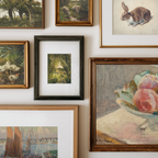 A gallery wall display of multiple framed art prints and paintings, including landscapes, still life fruit, a rabbit, ducks, and a harbor scene, arranged on a white wall.