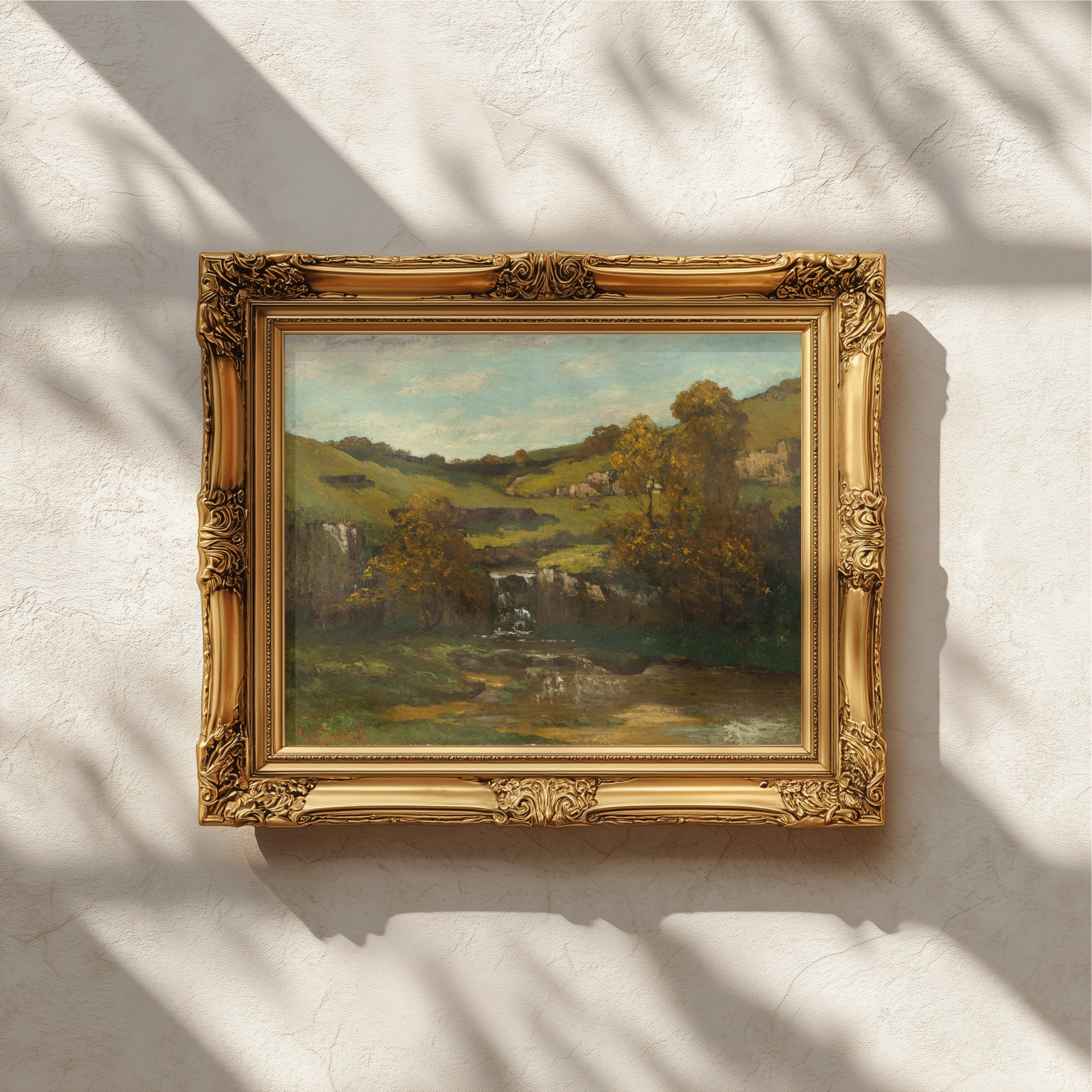 An ornate gold picture frame containing a landscape oil painting of rolling green hills, trees, and a stream, mounted on a textured white wall with soft leaf shadows cast across it.