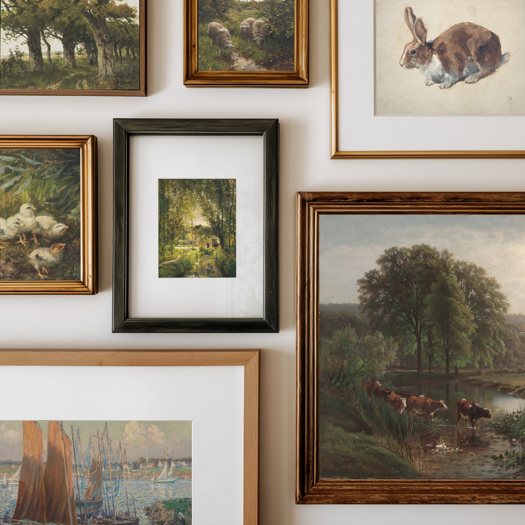 A gallery wall display of multiple framed art prints and paintings, including landscapes, a rabbit illustration, ducks, and cows by a river, arranged in a clean grid on a white wall.