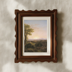 A framed landscape art print featuring a scenic valley with a tree and distant mountains under a soft sky, matted with white and housed in a dark brown wooden frame with a scalloped edge, mounted on a textured beige wall.